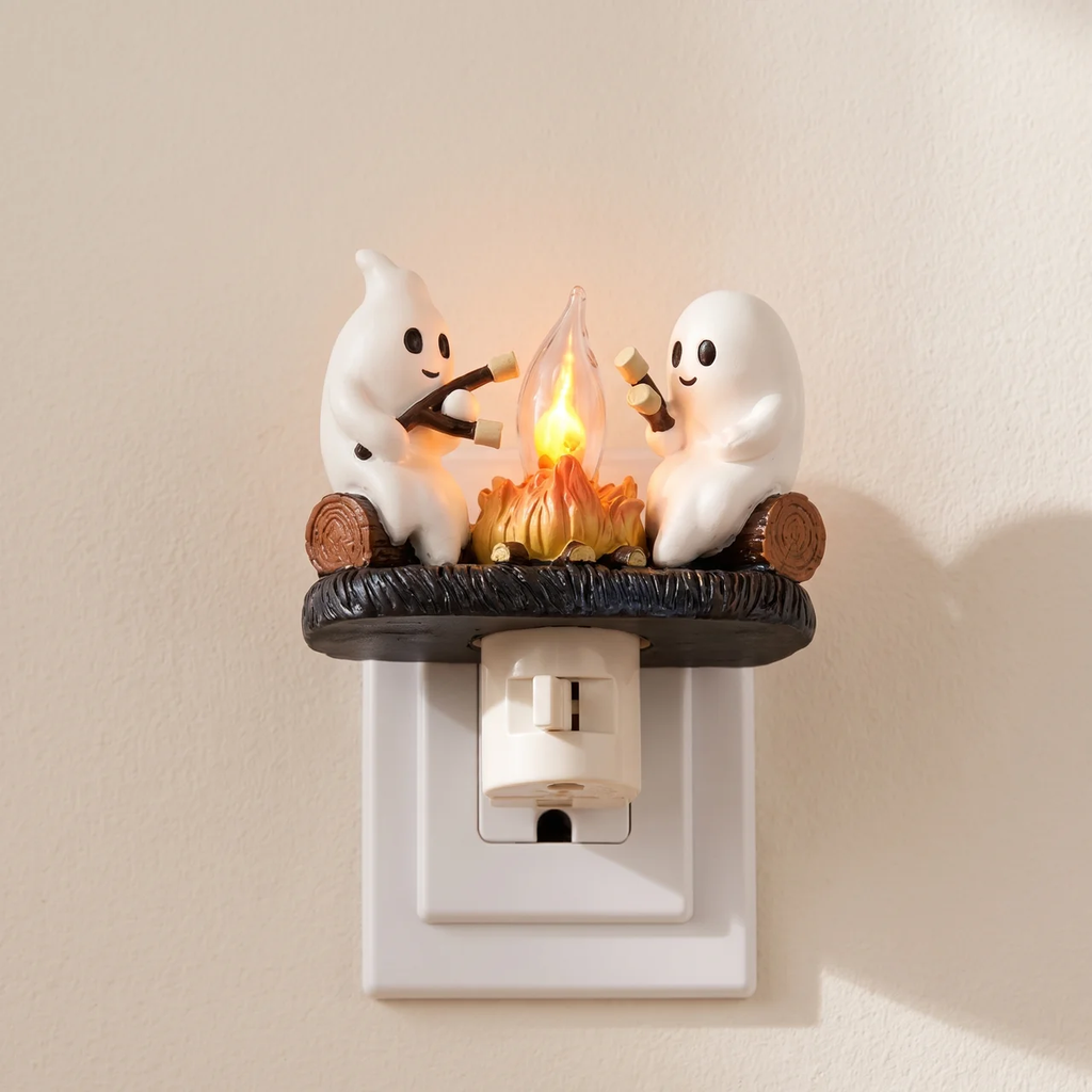 A ghost-themed night light plug-in featuring two white cartoon ghosts sitting around a glowing campfire on a dark base, mounted on a white wall outlet against a beige wall.