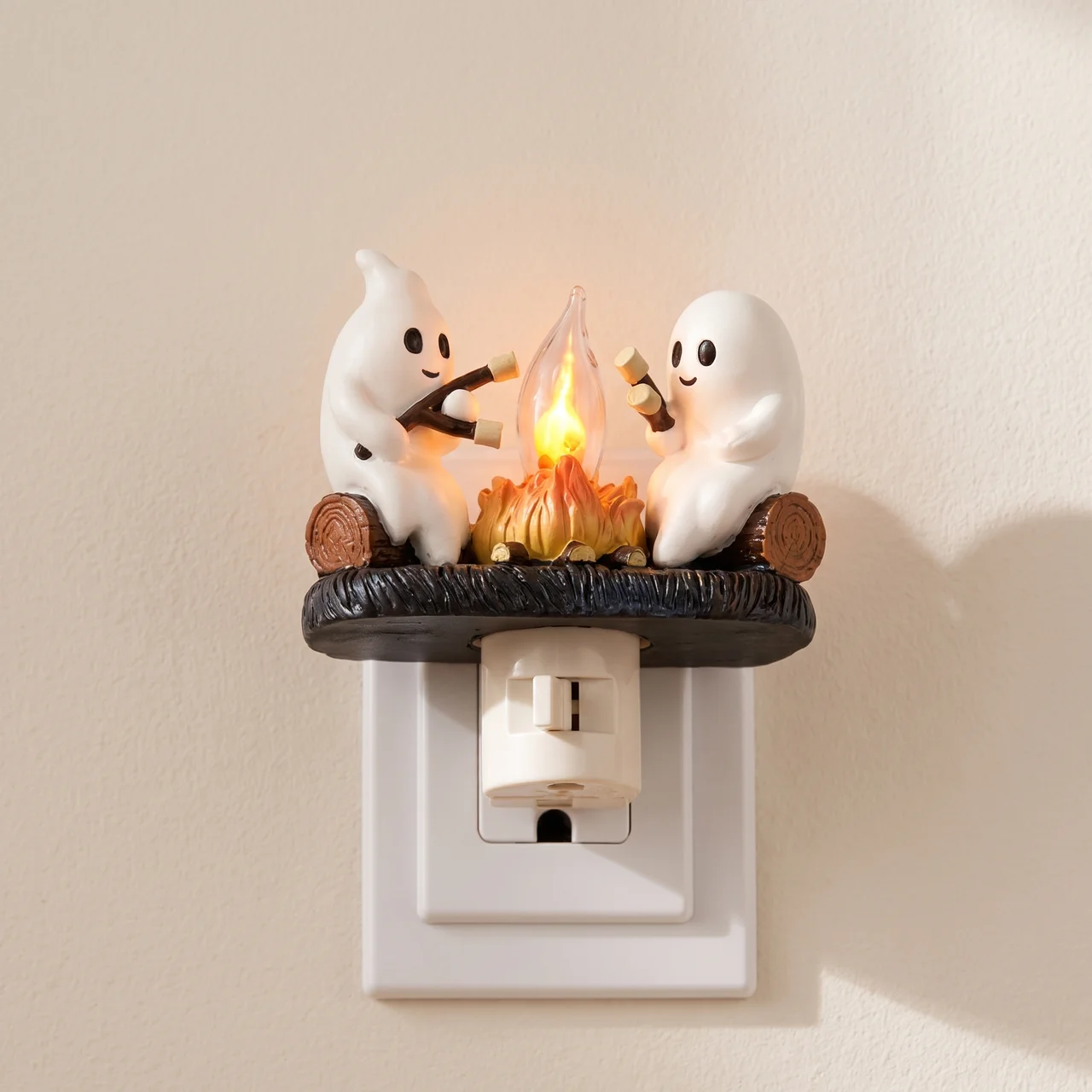 A ghost-themed night light plug-in featuring two white cartoon ghosts sitting around a glowing campfire on a dark base, mounted on a white wall outlet against a beige wall.