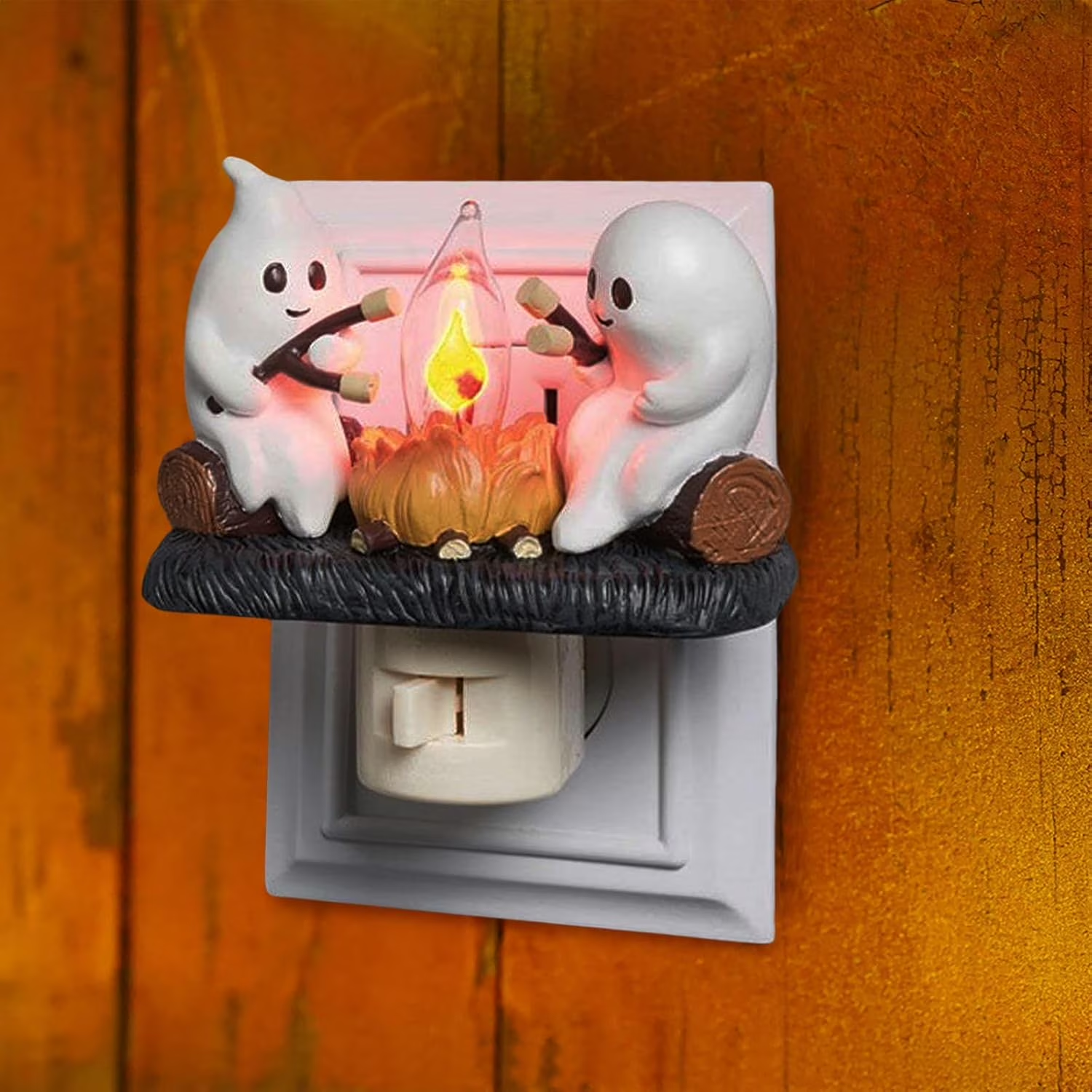 A Halloween-themed night light plug-in featuring two white cartoon ghosts roasting marshmallows around a glowing campfire on a dark textured base, shown plugged into a wall outlet against a warm wood-paneled background.