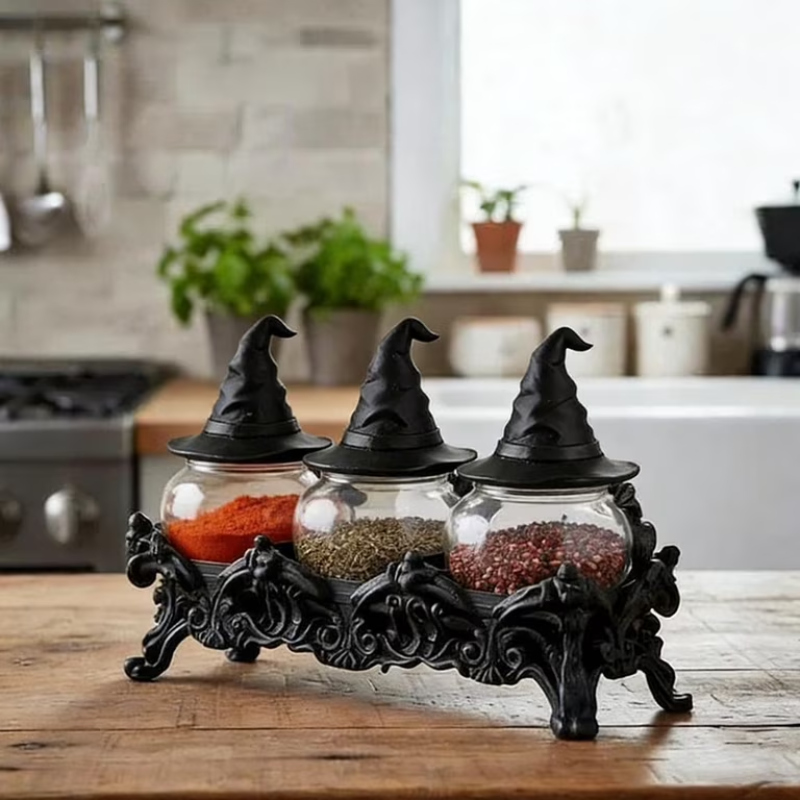 Three glass spice jars with pointed black witch hats are displayed on an ornate black metal base on a wooden table, with a blurred kitchen background.