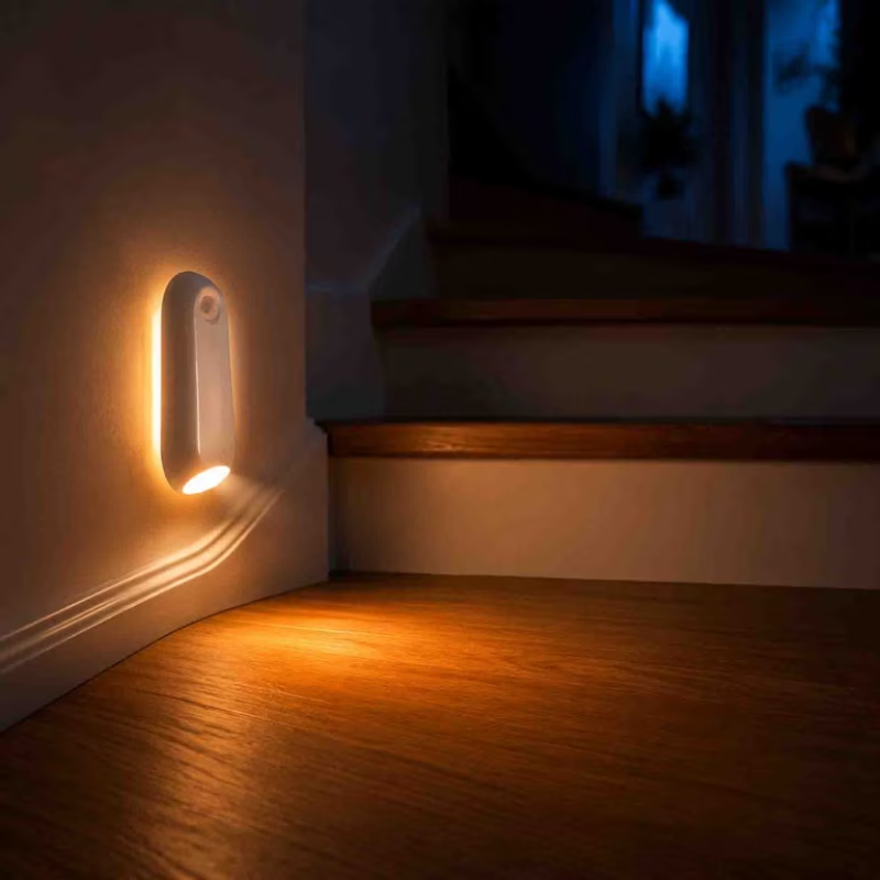 A small white wall-mounted LED night light with a warm yellow glow, shown lit on a staircase landing in a dimly lit home interior.
