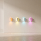 Four small cylindrical wall-mounted lights in different colors (orange, yellow, blue, pink) arranged in a row on a plain beige wall, casting soft light downwards.
