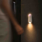 A white cylindrical wall-mounted light fixture with a glowing warm yellow light and a small purple indicator, mounted on a dark tiled wall. A blurred person walks past in the foreground.