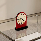 A modern arch-shaped desk clock with a red body, cream dial face, and black hands, displayed on a glass tabletop with a mesh surface.