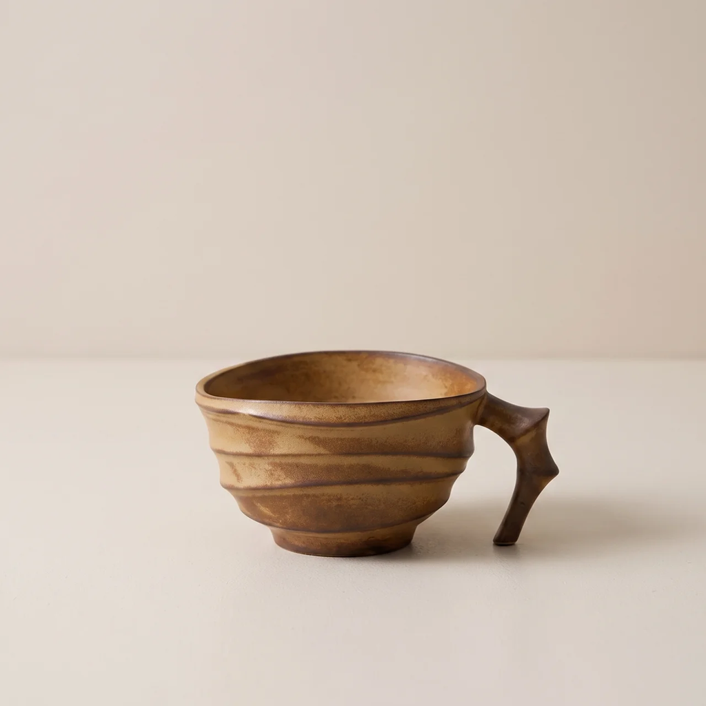 A handmade ceramic cup with a unique sculptural handle and a textured, ribbed exterior, shown on a neutral beige background.