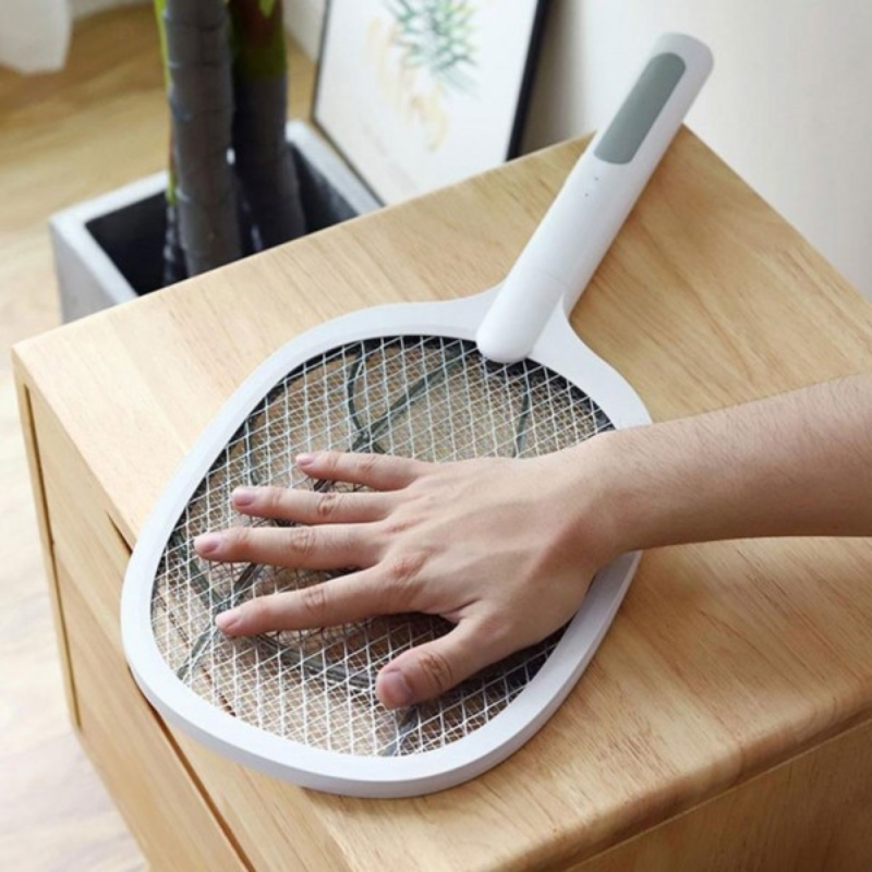 A white electric fly swatter with a wire mesh grid and a gray handle, shown with a person's hand resting on the mesh. The product is placed on a light-colored wooden table.