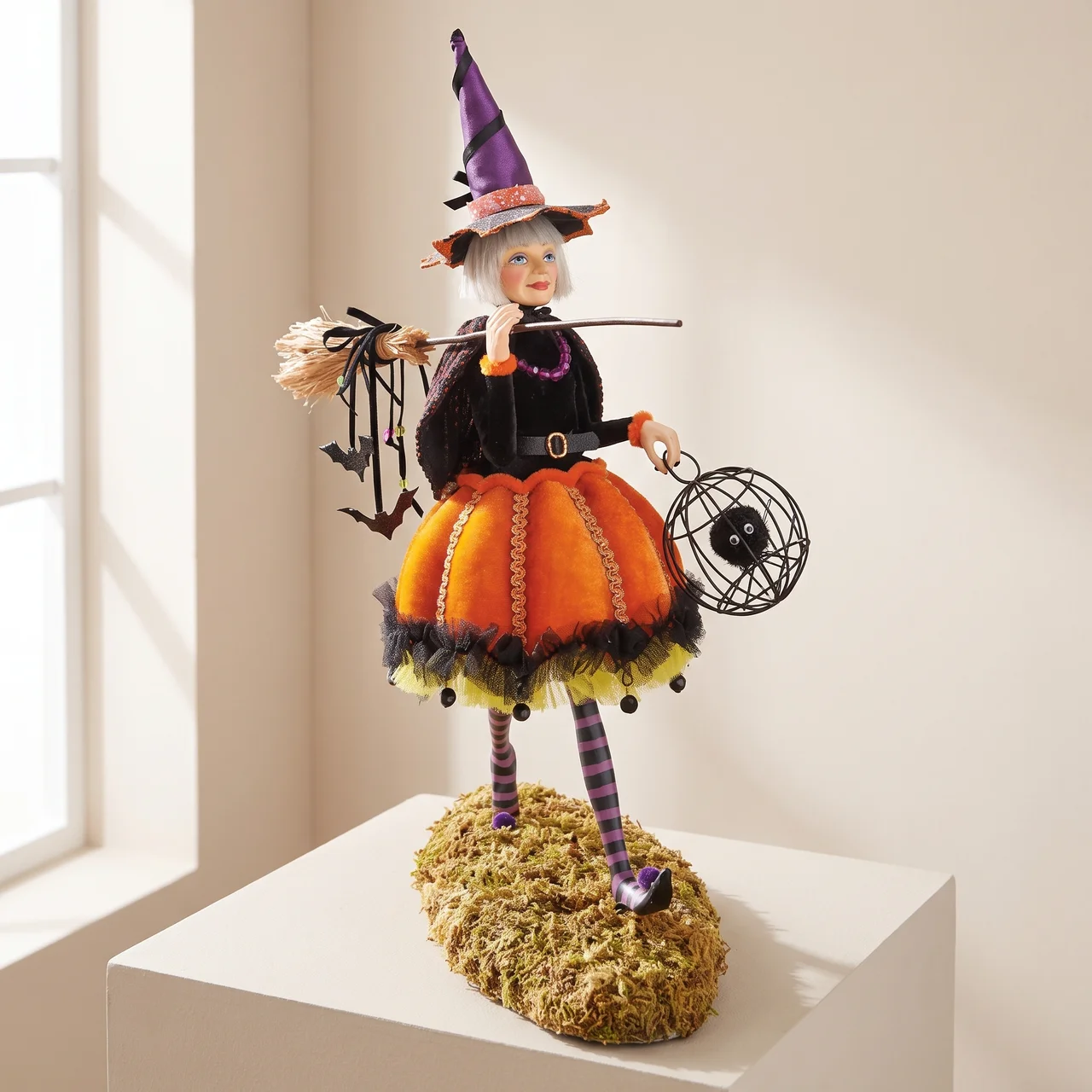 A decorative witch figurine stands on a mossy base in a bright indoor setting. The figurine has a purple witch hat, black dress, orange pumpkin skirt, striped purple-and-white stockings, and holds a broom and a spider web lantern.