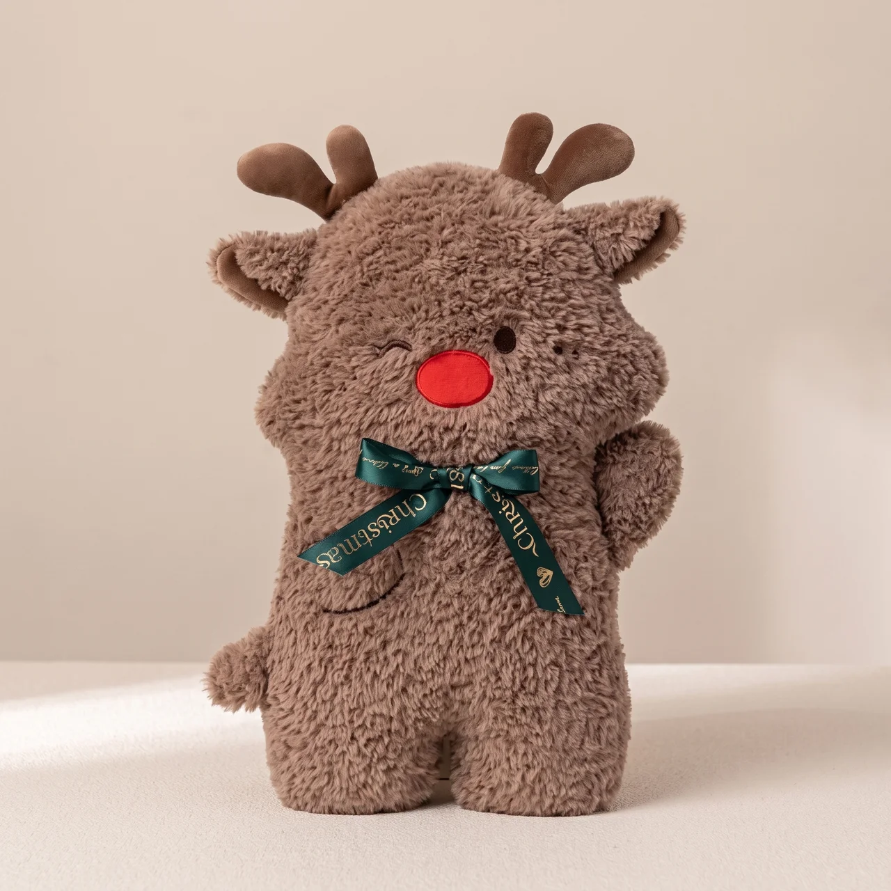 A brown plush reindeer toy with small antlers, a red nose, and a dark green ribbon bow around its neck, photographed against a plain beige background.