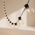 String lights with clear glass globe bulbs and black sockets, hanging against a warm beige wall with sunlight casting soft shadows.