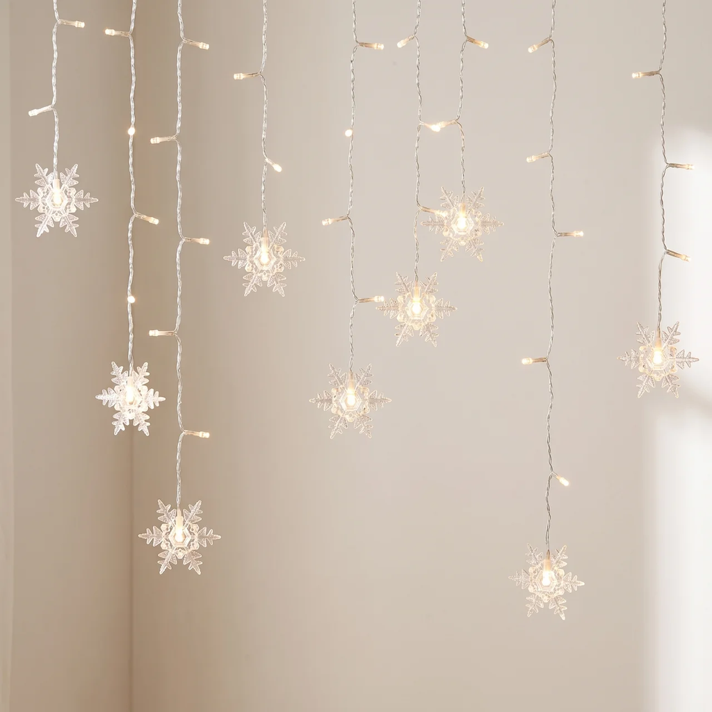 Warm white LED curtain string lights featuring hanging snowflake-shaped bulbs, shown illuminated against a plain beige wall.
