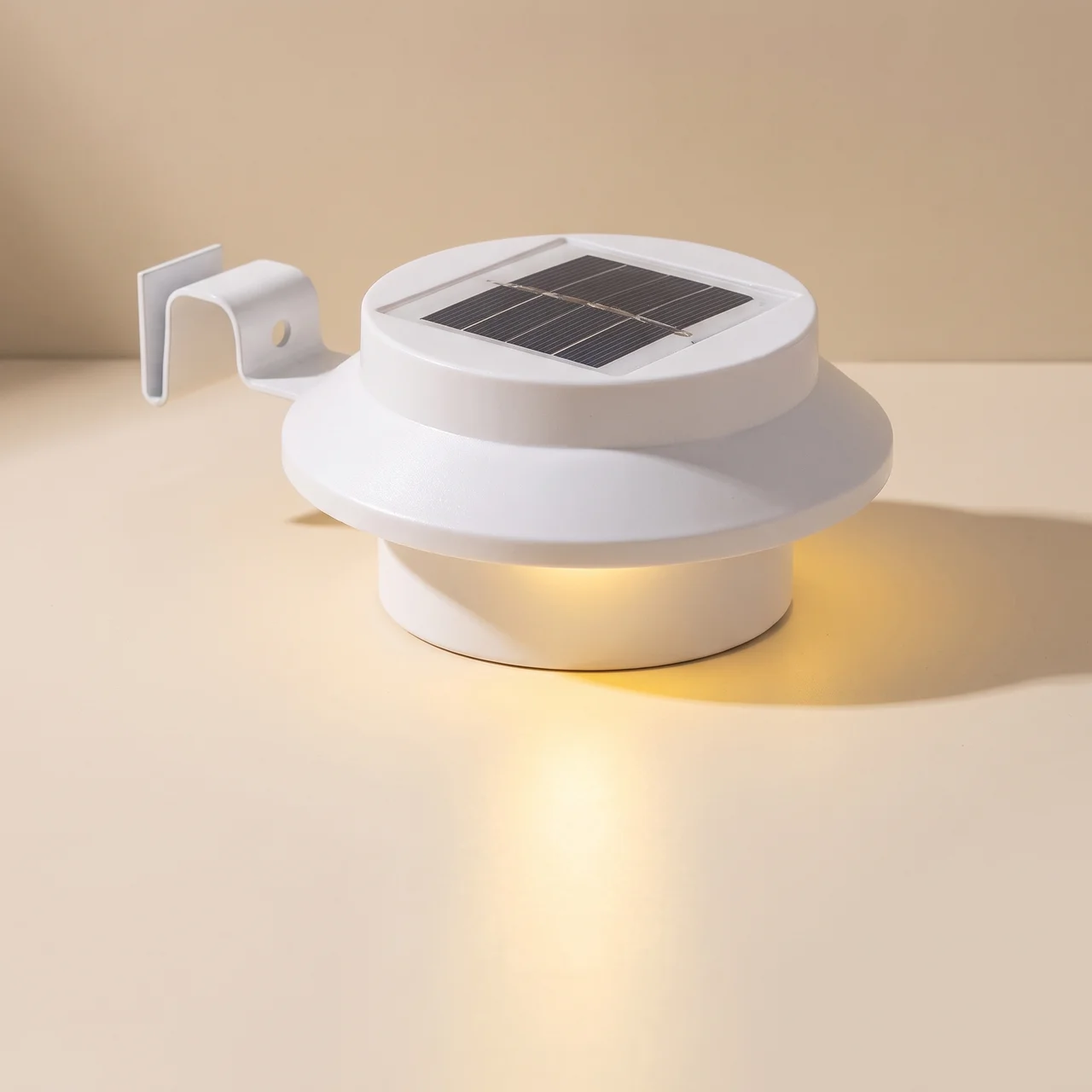 A white solar-powered outdoor light fixture with a circular top panel containing a dark solar cell and a side mounting bracket, shown lit on a warm beige background.
