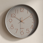 A minimalist round wall clock with a light gray face and frame, rose gold numerals and hands, and a subtle drop shadow on a textured off-white wall.