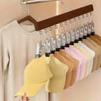 A wooden hanger with multiple metal clothes clips holds a row of baseball caps in different colors, including yellow, tan, brown, beige, pink, and white. A cream-colored sweater hangs on the left, and a hand is visible holding the front yellow cap.