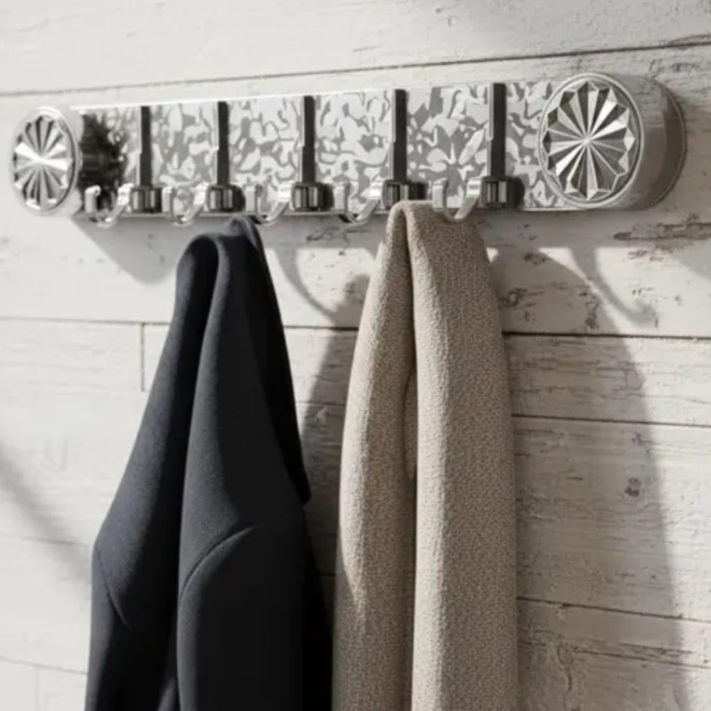 A wall-mounted silver coat rack with five hooks and decorative side caps, shown hanging a dark coat and a light beige towel against a whitewashed wood wall.