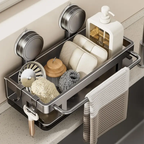 A stainless steel sink organizer tray mounted with two suction cups on a bathroom or kitchen counter. The tray holds soap bars, a white pump bottle, a sponge, a scrubber, and a hanging towel, with a faucet visible behind it.