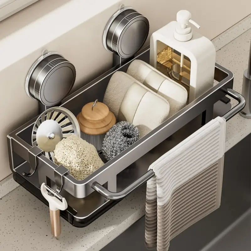 A stainless steel sink organizer tray mounted with two suction cups on a bathroom or kitchen counter. The tray holds soap bars, a white pump bottle, a sponge, a scrubber, and a hanging towel, with a faucet visible behind it.