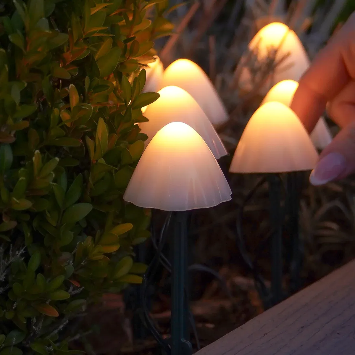 Mushroom-shaped outdoor string lights glowing warm white, shown planted among green shrubs with a human hand reaching toward one light.