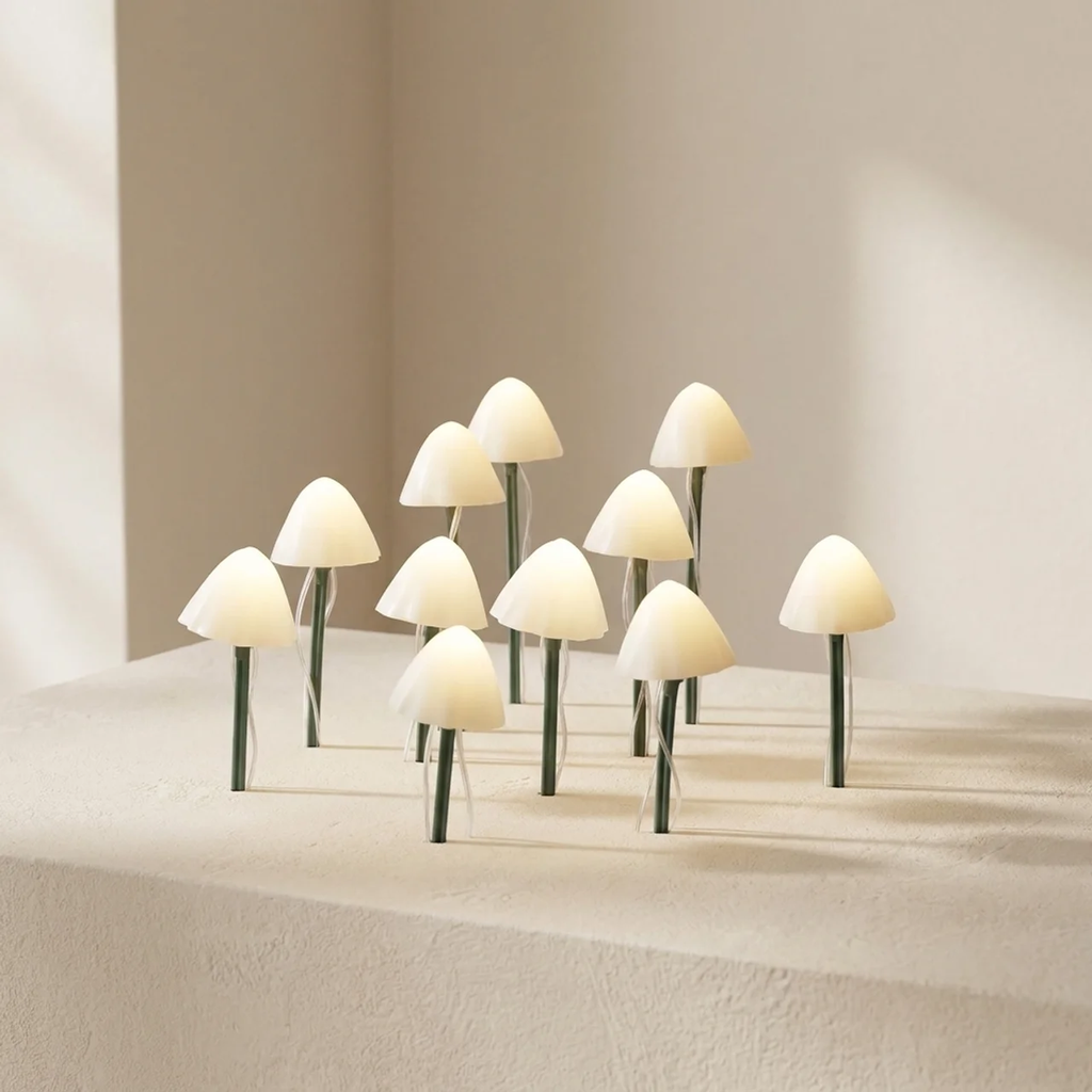 A set of small mushroom-shaped table lamps with glowing warm white caps and dark green stems, arranged on a textured beige surface against a neutral cream background.