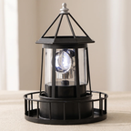 A black metal lantern-style table lamp with clear glass panels and a visible illuminated bulb, shown on a light beige surface against a soft neutral background.