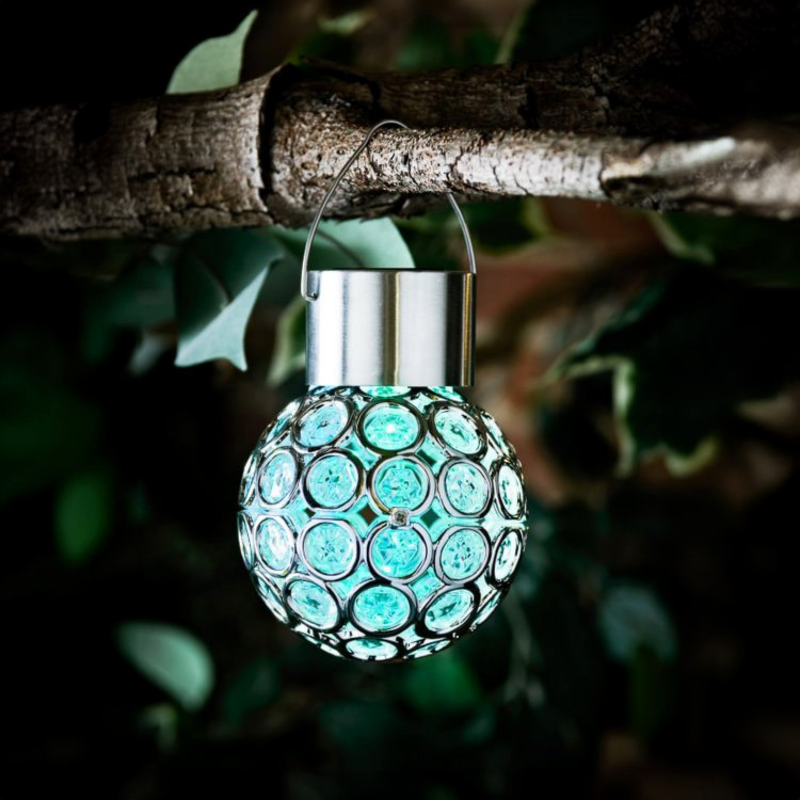 A hanging decorative light bulb ornament with a silver metal cap and handle, covered in circular turquoise-blue glass or acrylic discs, suspended from a tree branch outdoors against a dark leafy background.