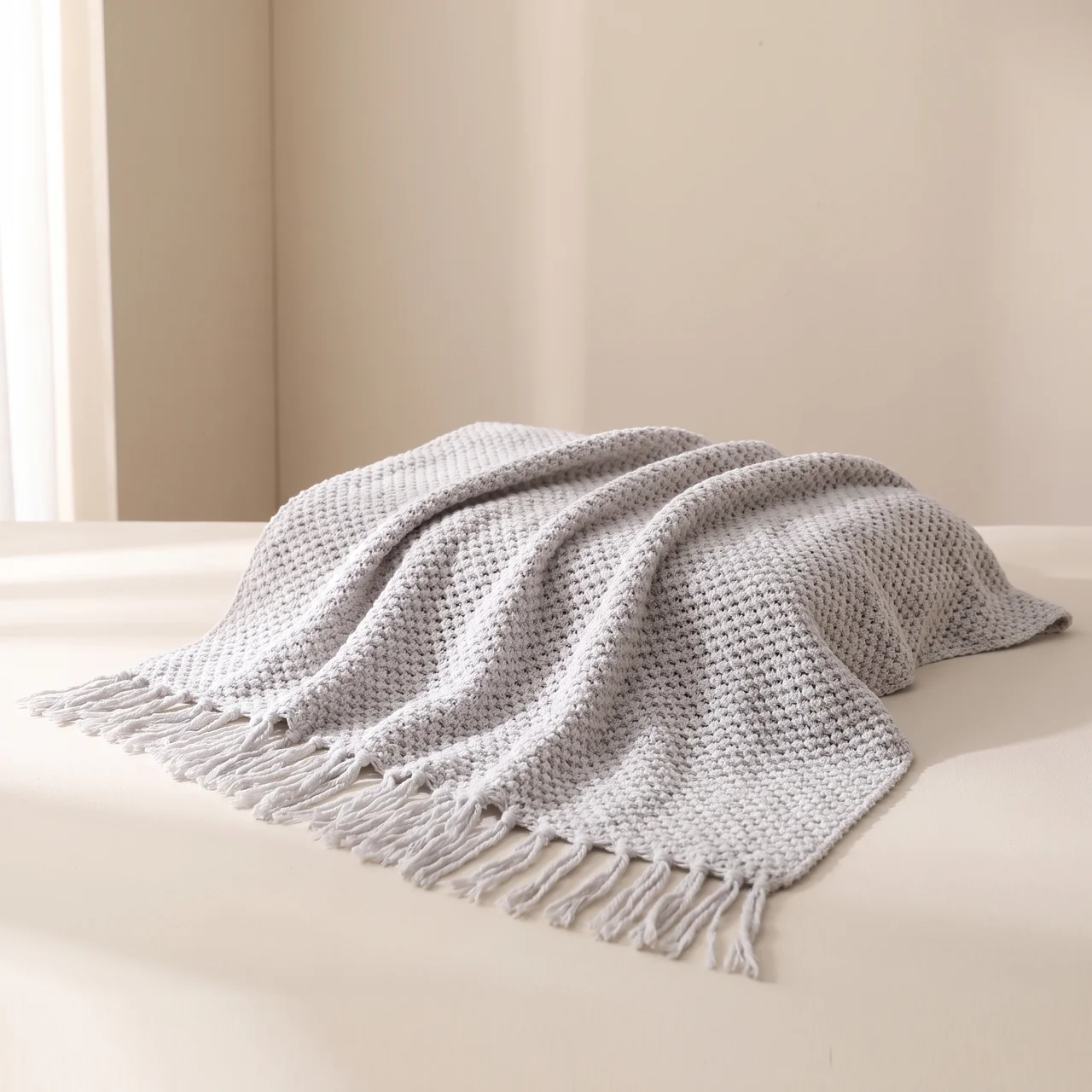 A light gray textured knit throw blanket with fringed edges, draped over a cream-colored surface against a neutral beige background.