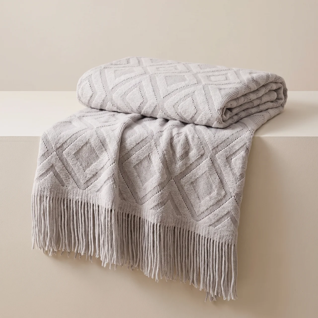 A folded and draped knit throw blanket in a light warm gray or greige tone, featuring a raised geometric diamond pattern and long fringe along one edge, displayed on a matching pedestal against a neutral beige background.