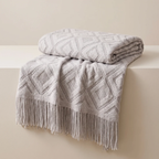 A folded and draped knit throw blanket in a light warm gray or greige tone, featuring a raised geometric diamond pattern and long fringe along one edge, displayed on a matching pedestal against a neutral beige background.
