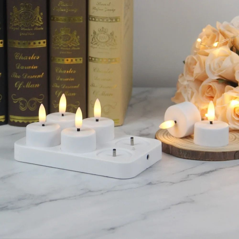 A white multi-candle plug-in holder with several small white tea light candles lit, styled on a marble surface with books and a rose arrangement in the background.