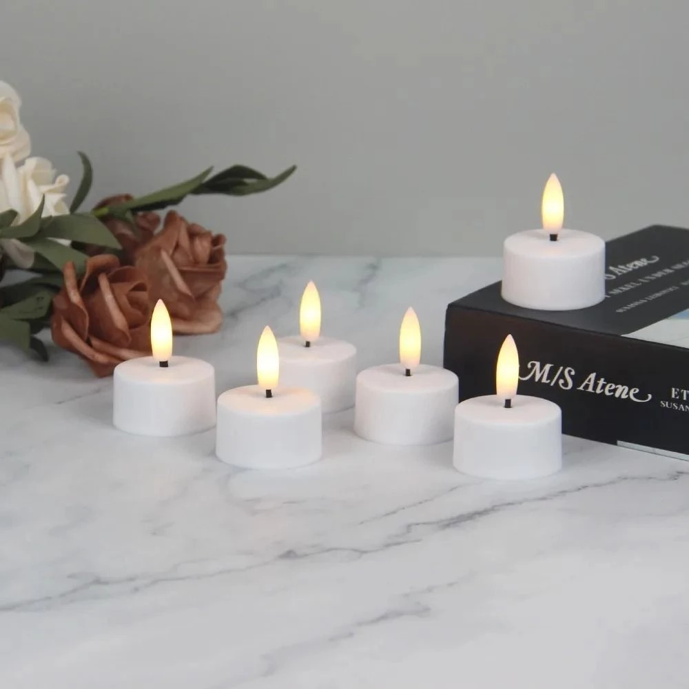 Set of six white candles with flames on a marble surface with a book and flowers in the background.