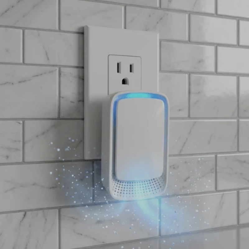 A white electronic plug-in device with a glowing blue light, shown plugged into a wall outlet against a white subway tile background.