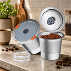 A stainless steel coffee capsule filling machine shown on a countertop with coffee beans, a cup of coffee, and a tiled kitchen background. The machine is open, showing a filled capsule and a loose powder chamber, with two clear plastic rings on the counter.