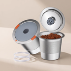 A stainless steel coffee capsule filling machine shown disassembled with its lid, inner capsule, and clear plastic seal on a warm beige background.