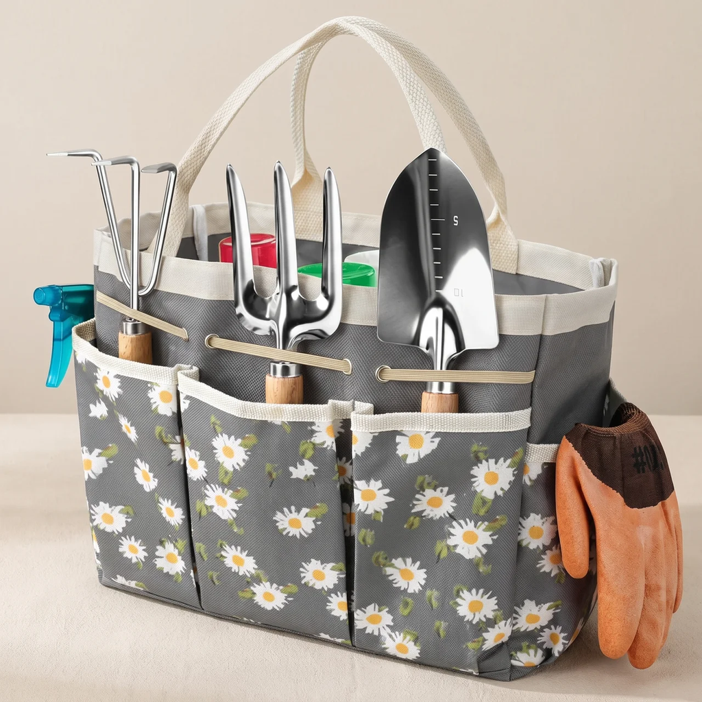 A gray fabric gardening tool organizer tote bag with a daisy floral print, holding various metal gardening tools, with a pair of orange gardening gloves hanging from the side, set against a neutral beige background.