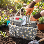 A gray fabric gardening tote bag with a white daisy print, shown in a garden with hands placing gardening tools inside. The bag has multiple front pockets and handles, with tools including a trowel, hand fork, and small pots visible.