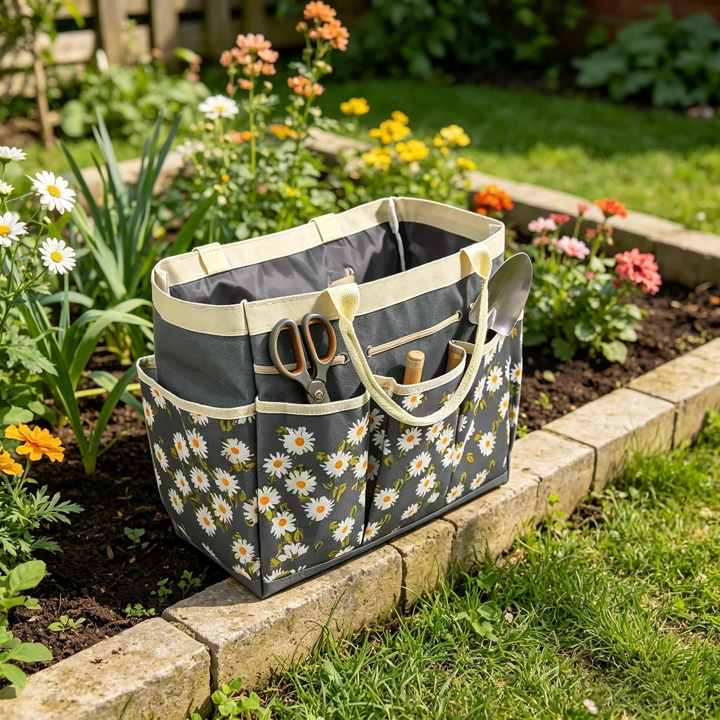 A gray fabric gardening tote bag with a white daisy floral print, beige handles, and storage pockets, shown sitting on a stone garden bed with flowers and grass in the background.