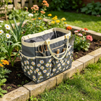 A gray fabric gardening tote bag with a white daisy floral print, beige handles, and storage pockets, shown sitting on a stone garden bed with flowers and grass in the background.
