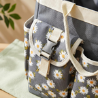 Close-up of a gray fabric tote bag with a daisy floral print and cream canvas straps, shown on a light surface with a soft green cloth and blurred leaves in the background.