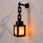 A black metal wall sconce with a geometric block design and a warm glowing orange light, mounted on a white marble wall.