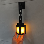 A black modern wall sconce with a glowing warm yellow light, held by a hand against a light gray tiled wall.