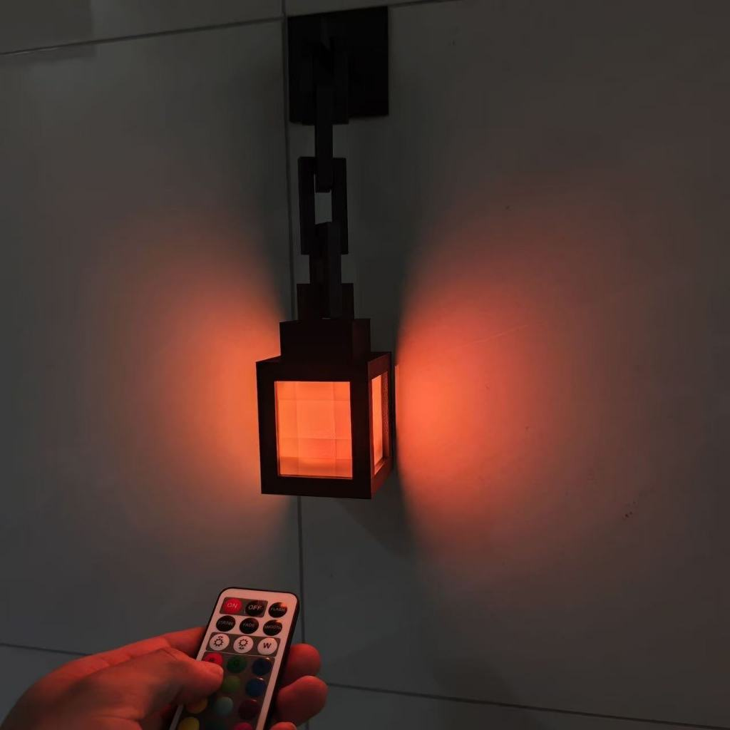 A black wall-mounted lantern-style light fixture with a glowing orange-red light, shown mounted on a gray wall. A hand in the lower left holds a small remote control.