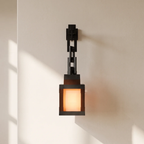 A modern black metal wall sconce with a glowing warm orange light, mounted on a beige wall with soft window shadow patterns.