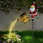 A festive garden stake decoration featuring a Santa Claus figure holding a glowing decorative watering can. The watering can casts a warm yellow light, simulating water, which drips onto flowers in a garden bed.