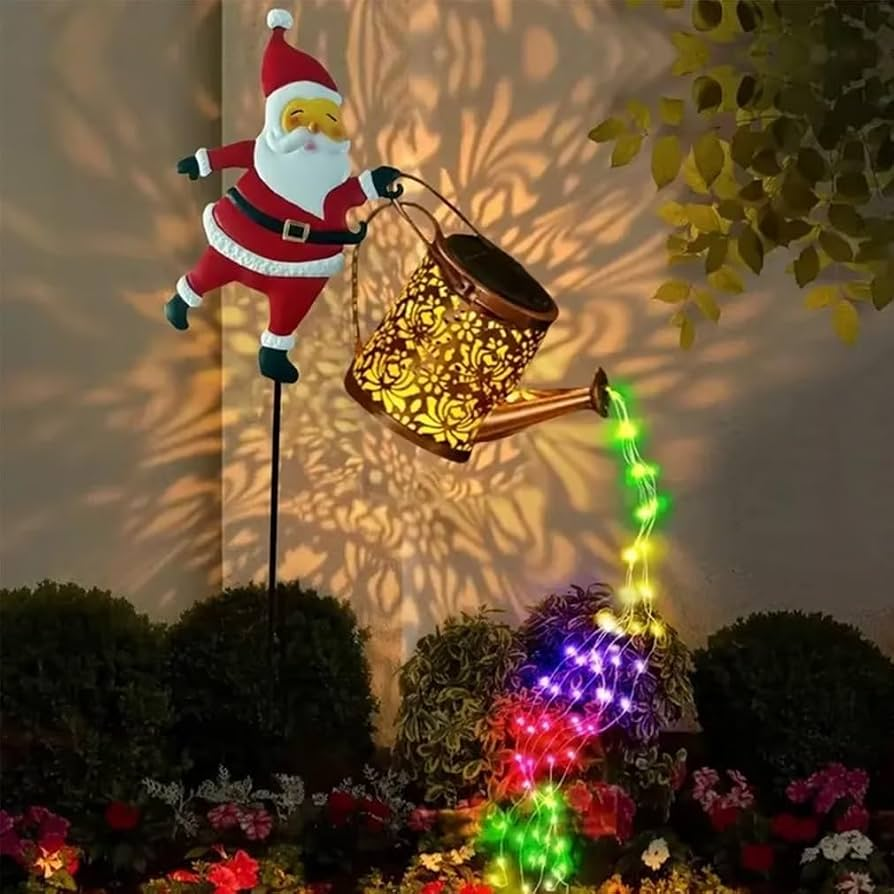A festive garden decoration featuring a Santa Claus figure holding a glowing gold watering can that casts colorful light patterns on a wall above a flower bed.