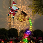 A festive garden decoration featuring a Santa Claus figure holding a glowing gold watering can that casts colorful light patterns on a wall above a flower bed.
