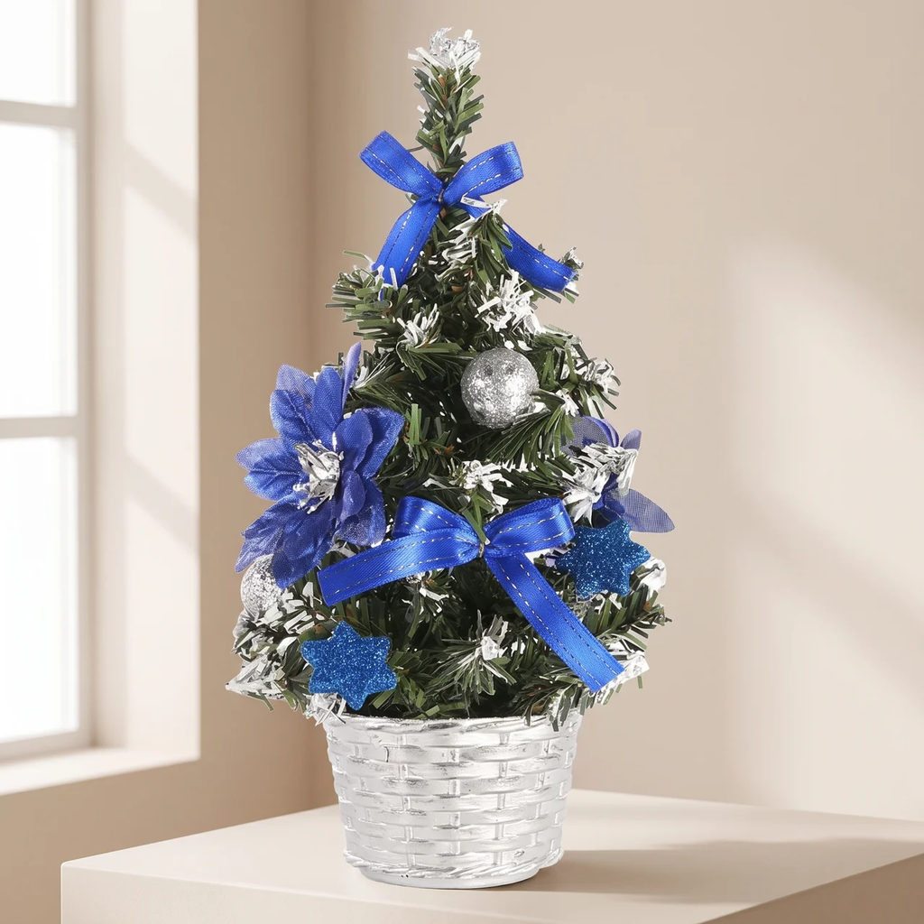 A small artificial Christmas tree in a clear woven basket, decorated with blue ribbons, blue flowers, silver ornaments, and white snowflake accents, set on a table in front of a softly lit beige wall.