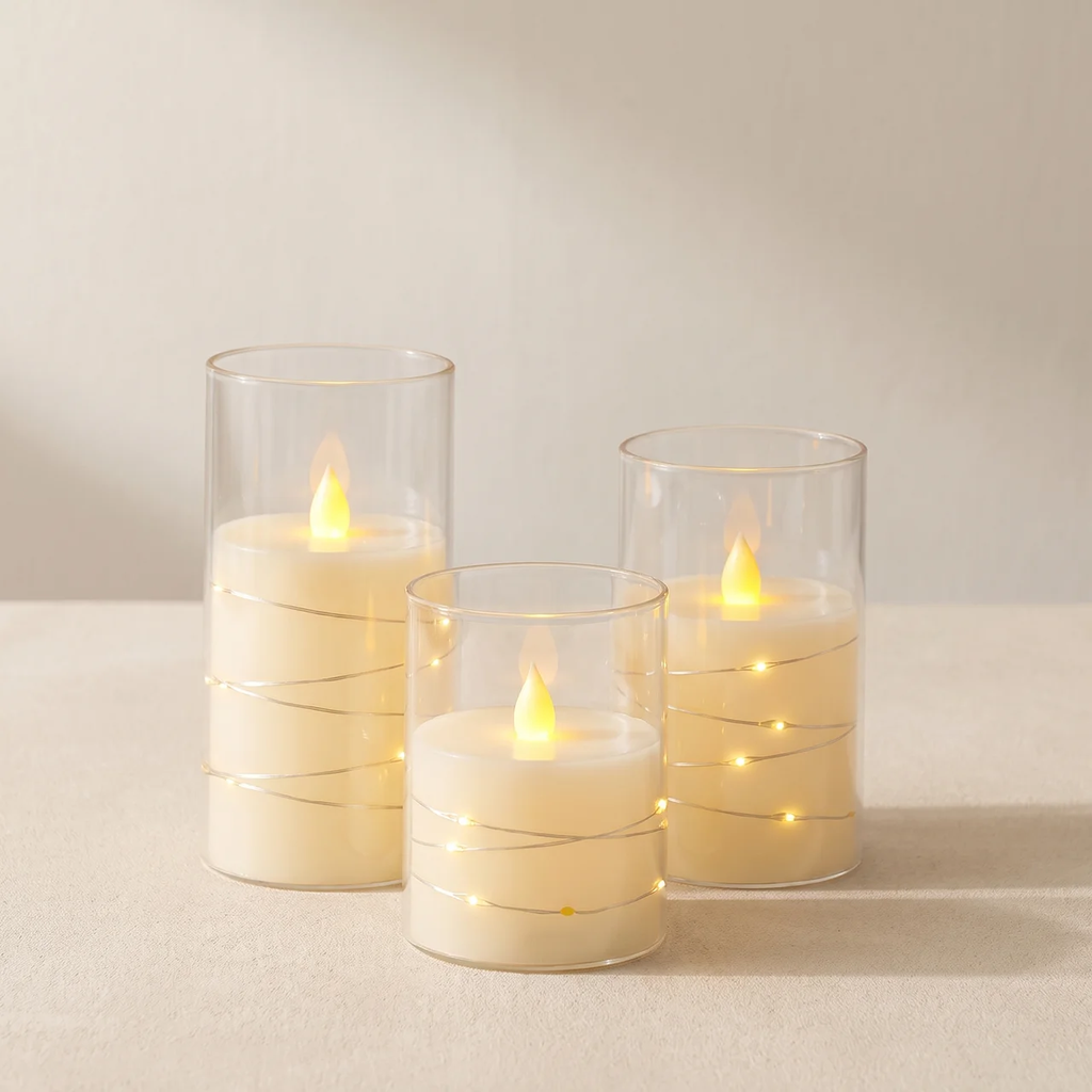 Three glass candle holders with thin gold wire accents, each containing a lit cream-colored candle, arranged on a neutral beige surface against a soft beige background.