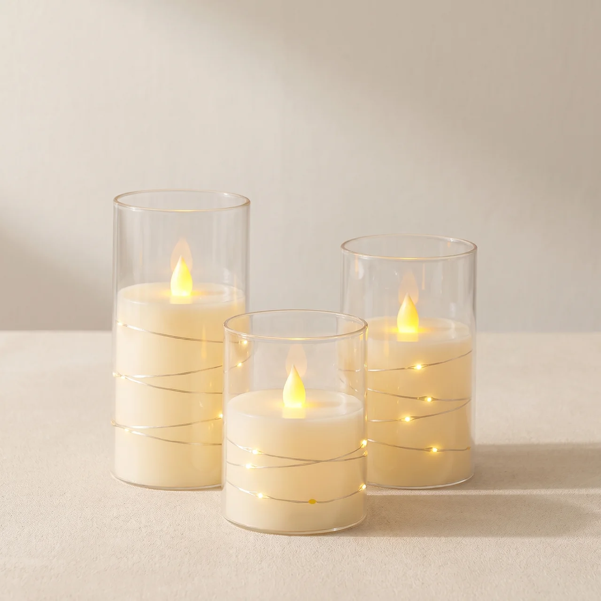 Three glass candle holders with thin gold wire accents, each containing a lit cream-colored candle, arranged on a neutral beige surface against a soft beige background.