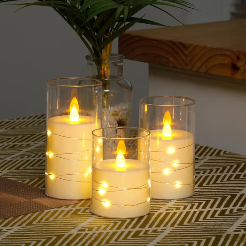 Three lit candles in glass holders on a patterned surface with a plant in the background