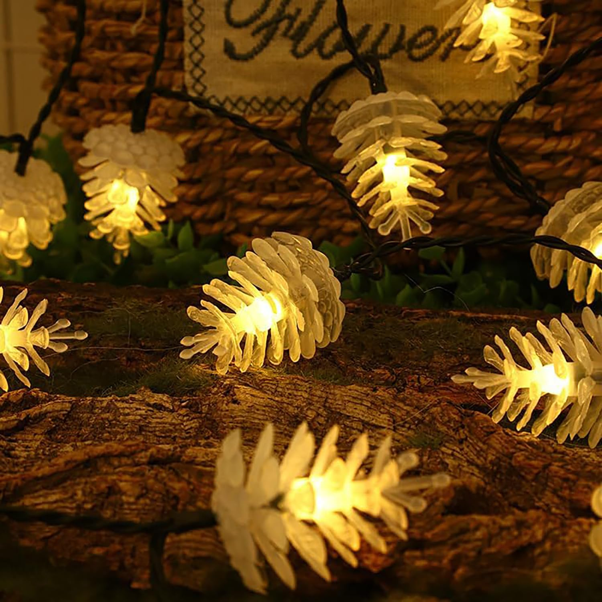 Pinecone-shaped string lights glowing with warm yellow light, draped over a rustic wooden log with green foliage and a woven basket in the background.