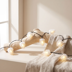 Warm white LED pinecone string lights draped over a rustic wood log and beige linen fabric in a softly lit interior setting.